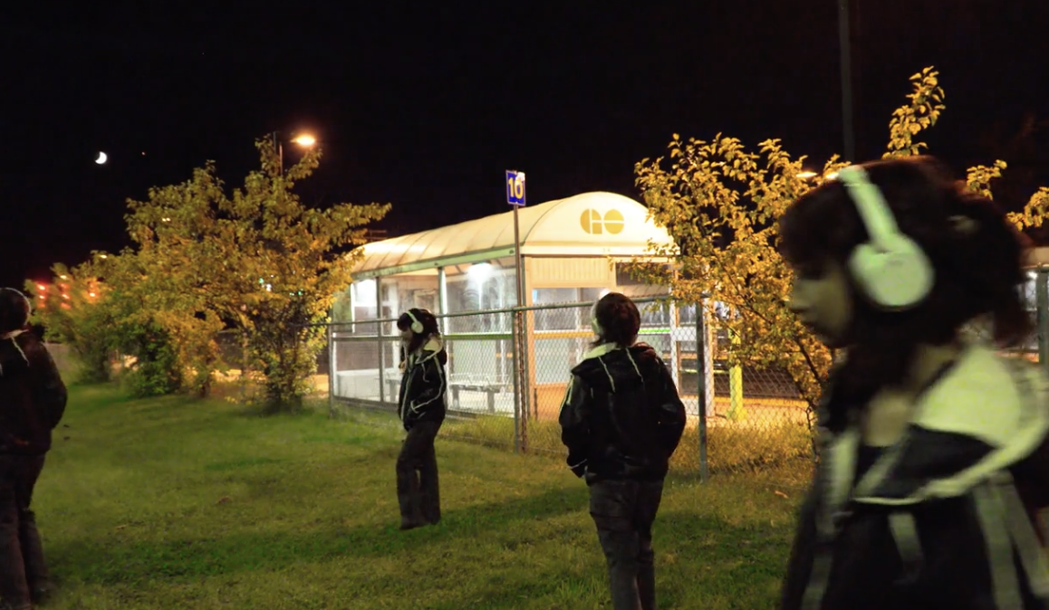 Night exterior with headphone-wearing figures near a transit shelter